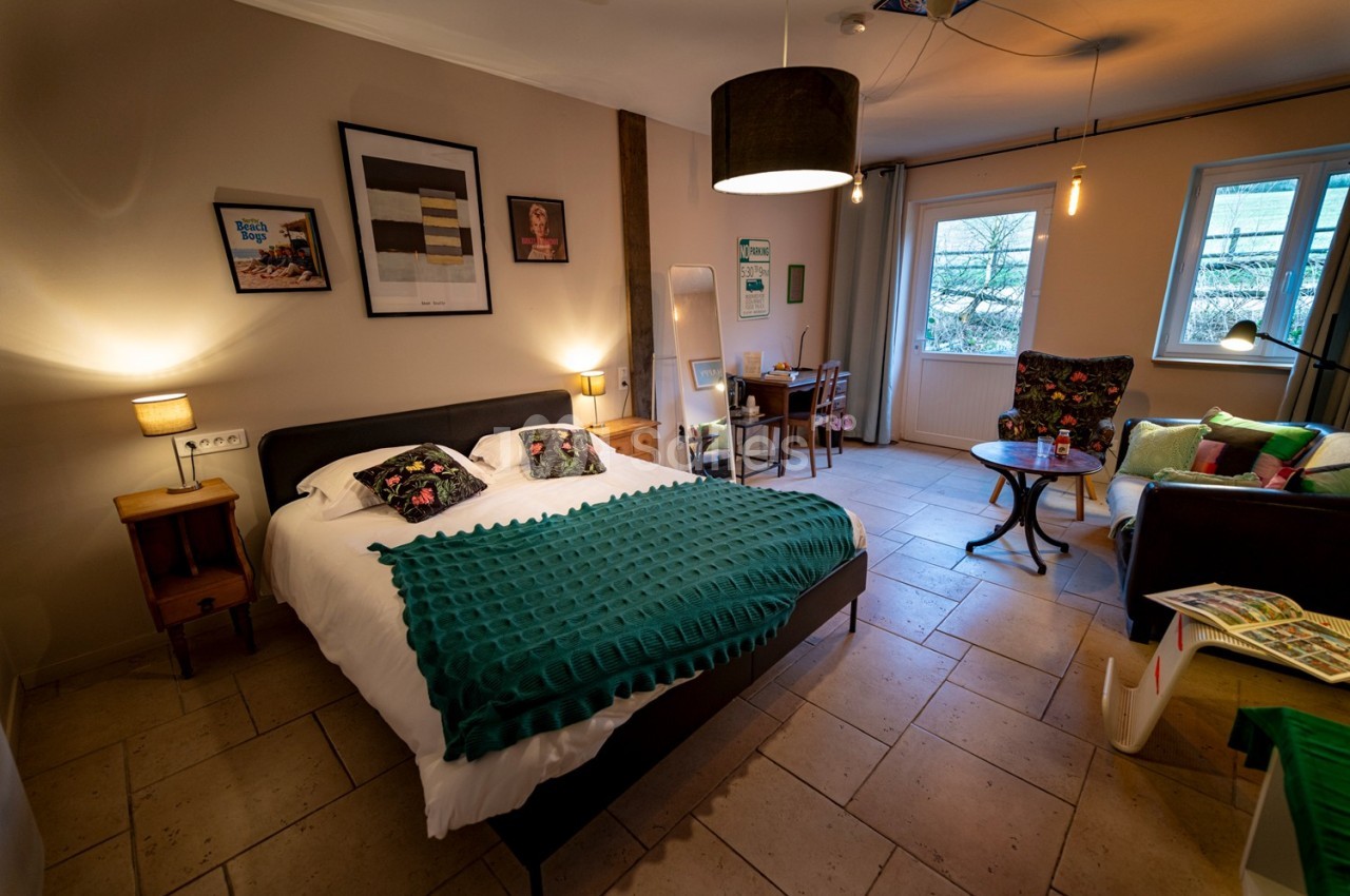 Chambre lumineuse avec lit double, coin salon, bureau, décoration murale et vue sur un jardin à travers les fenêtres.