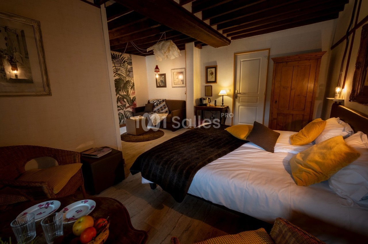 Chambre chaleureuse avec lit double, coussins jaunes, mobilier en bois, coin salon et éclairage tamisé.