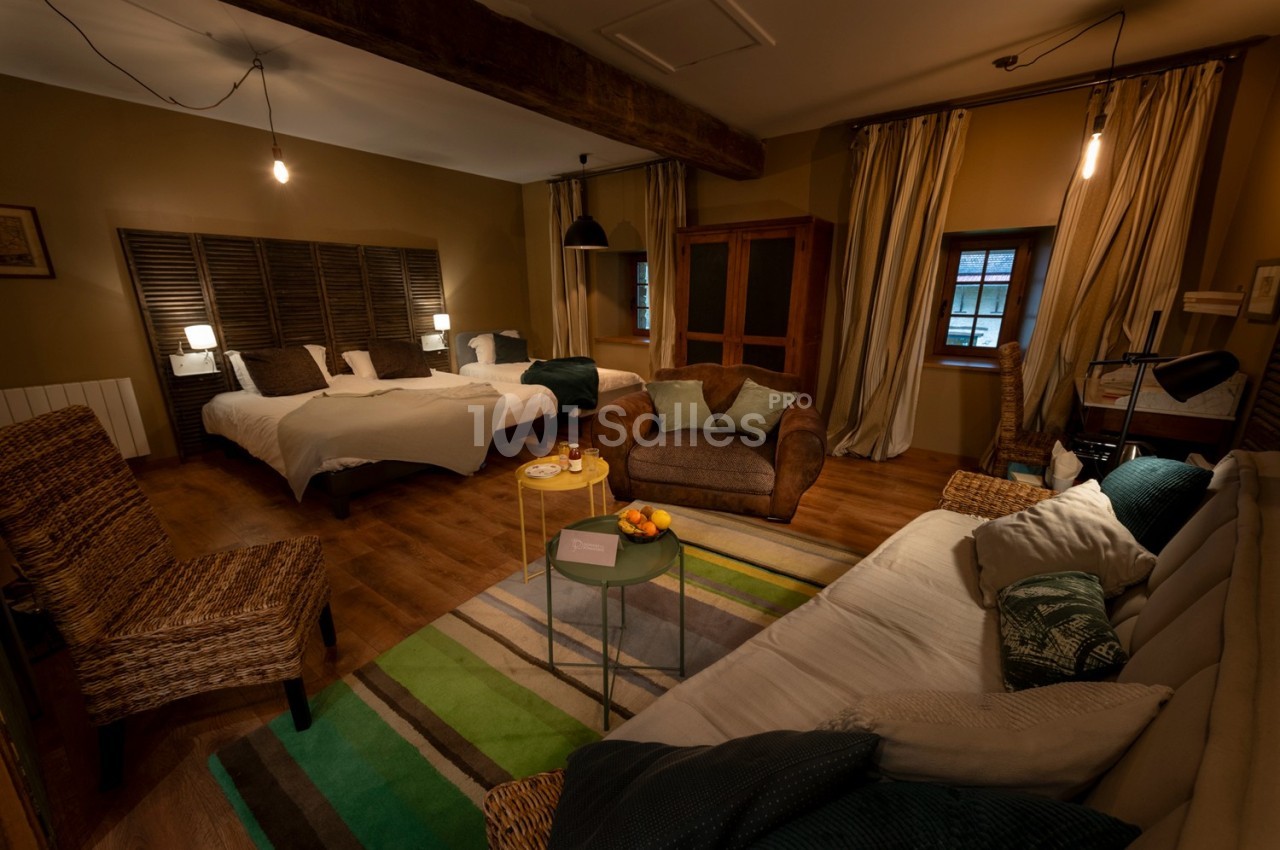 Chambre chaleureuse avec lits, canapé, fauteuils en osier, table basse et éclairage tamisé, décor rustique.