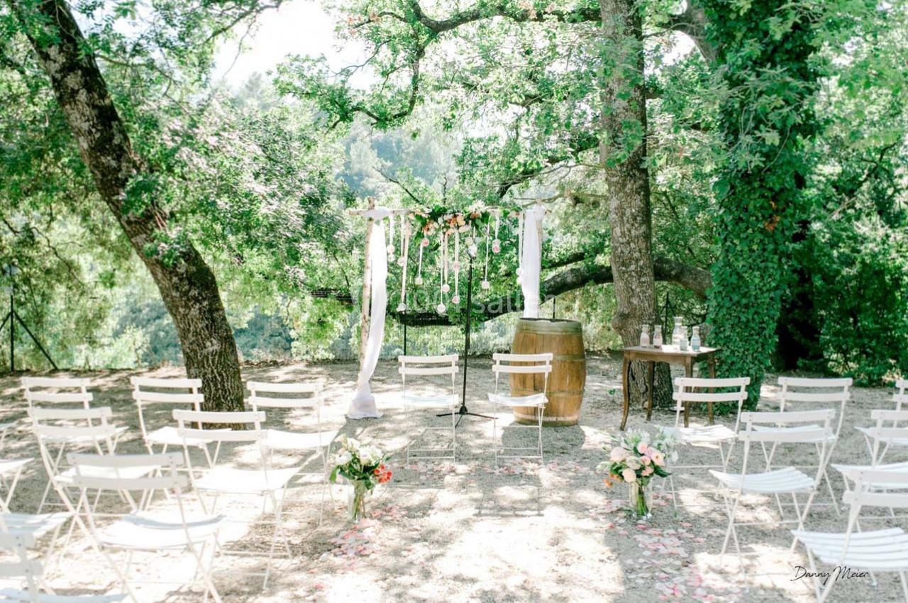 Location salle Signes (Var) - Château de Cancerilles #17 Chaises blanches disposées devant une arche décorée de fleurs et de voilages, dans un cadre boisé en plein air.
