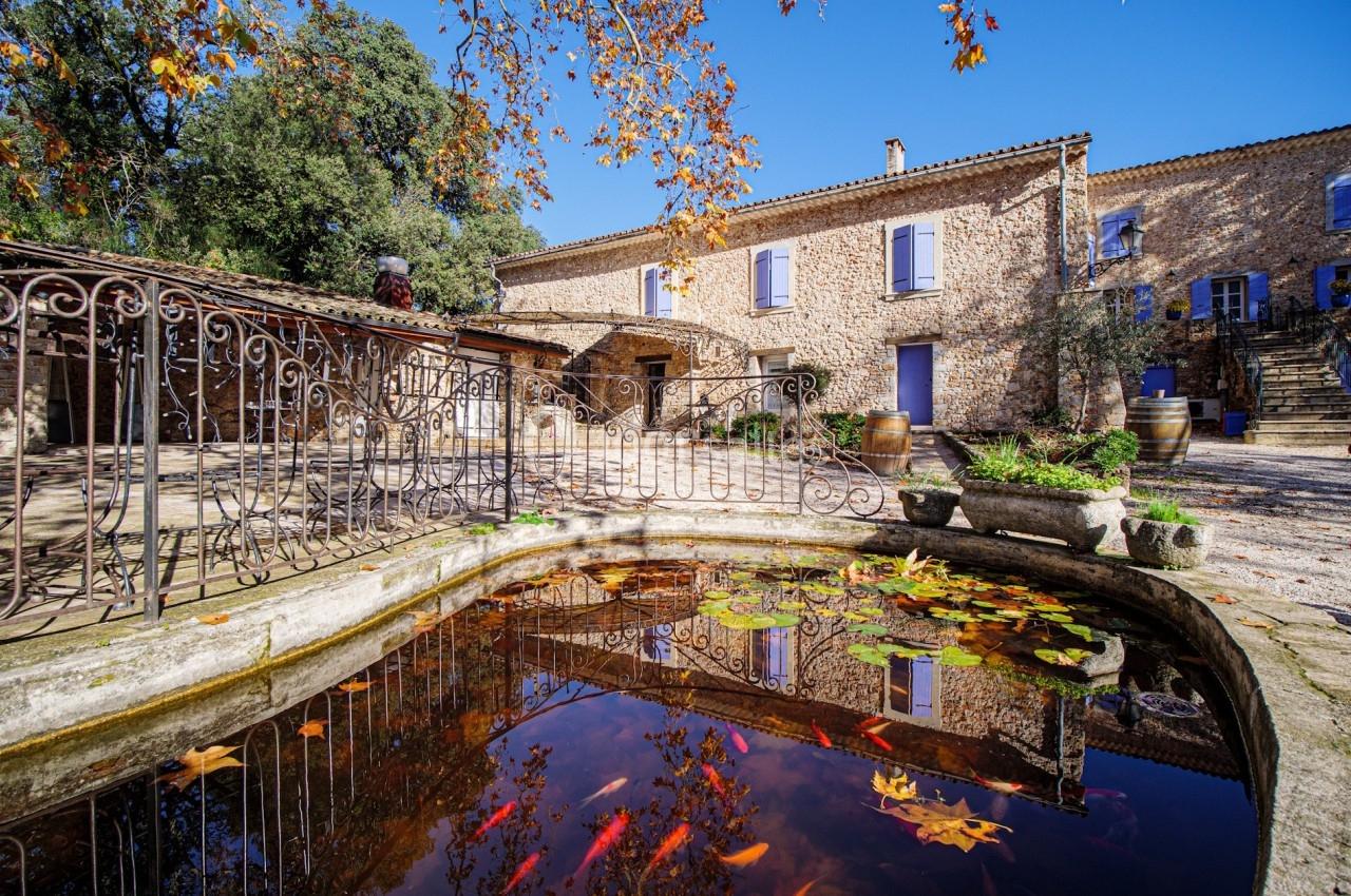 Location salle Signes (Var) - Château de Cancerilles #2 Bassin avec des poissons rouges entouré de feuilles d'automne, devant une bâtisse en pierre sous un ciel dégagé.
