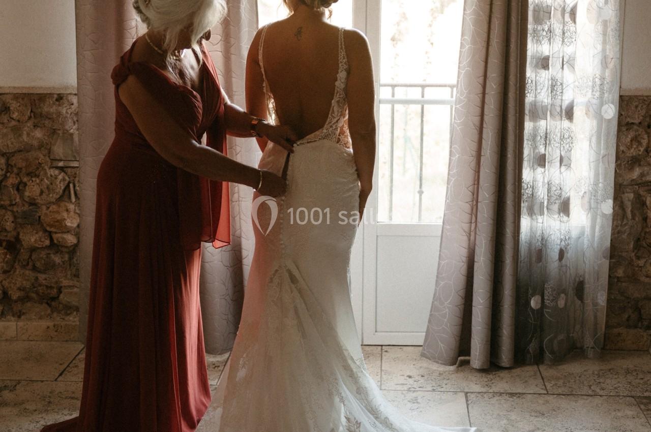 Location salle Signes (Var) - Château de Cancerilles #37 Une femme aide une mariée à ajuster sa robe dans une pièce lumineuse avec des rideaux et une porte-fenêtre en arrière-plan.