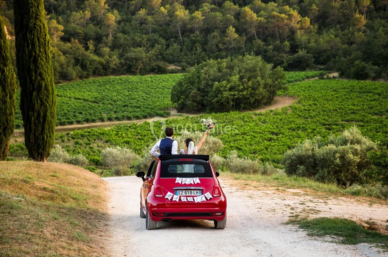 Location salle Signes (Var) - Château de Cancerilles #35 Un couple en tenue de mariage dans une voiture rouge décapotable sur un chemin entouré de vignes et de verdure.