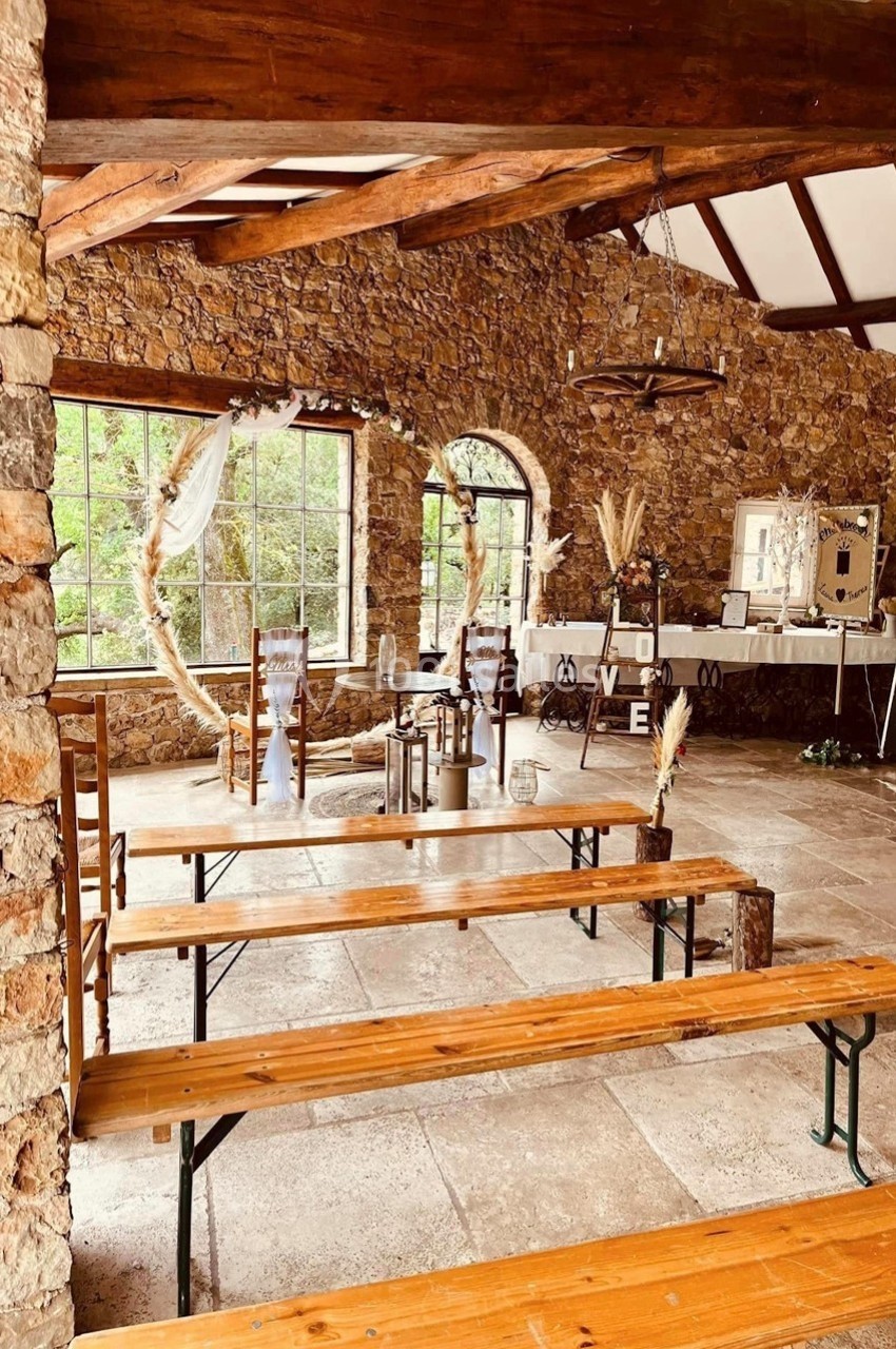 Location salle Signes (Var) - Château de Cancerilles #8 Salle en pierre avec bancs en bois, décor champêtre, grandes fenêtres et table ornée pour un événement.