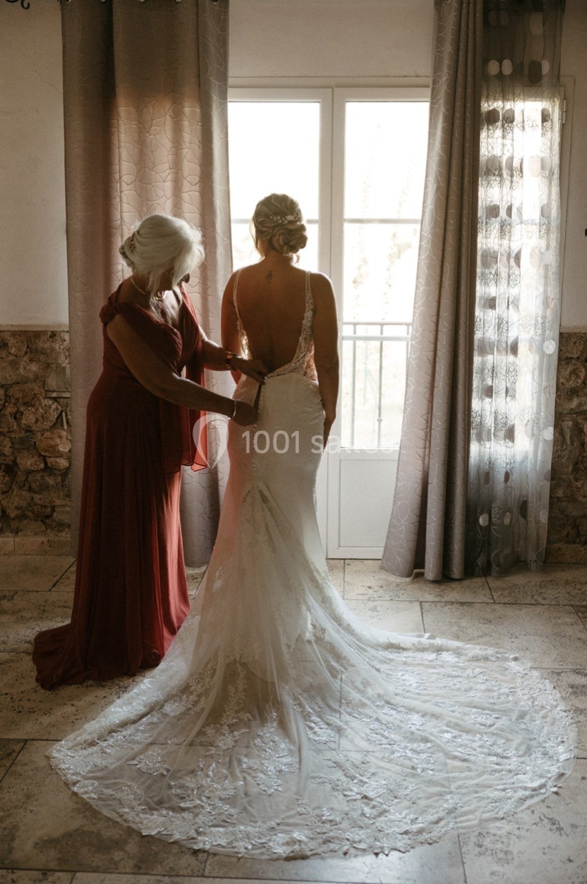 Location salle Signes (Var) - Château de Cancerilles #56 Une femme aide une mariée à ajuster sa robe dans une pièce lumineuse près d'une fenêtre.