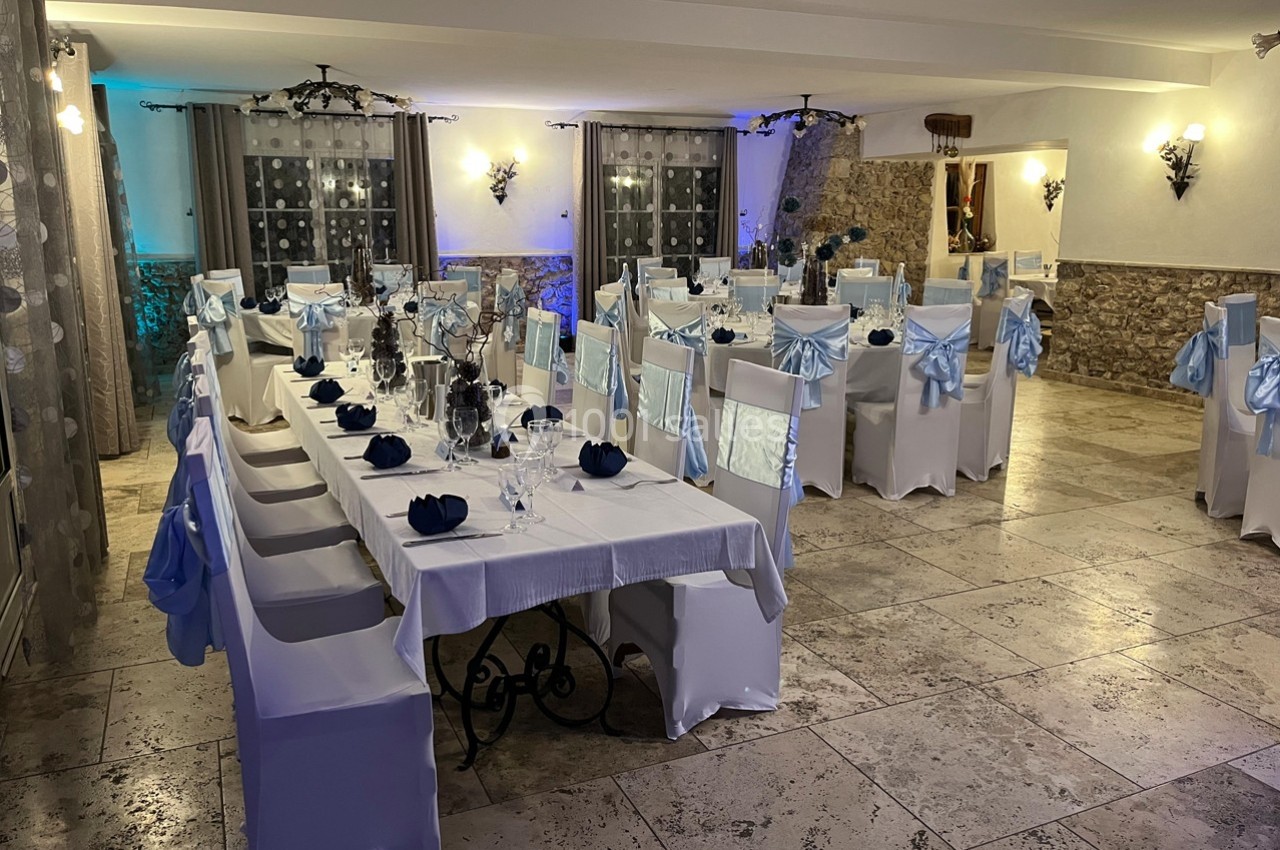 Location salle Signes (Var) - Château de Cancerilles #9 Salle de réception décorée avec des tables dressées, des chaises blanches ornées de nœuds bleus et un éclairage chaleureux.