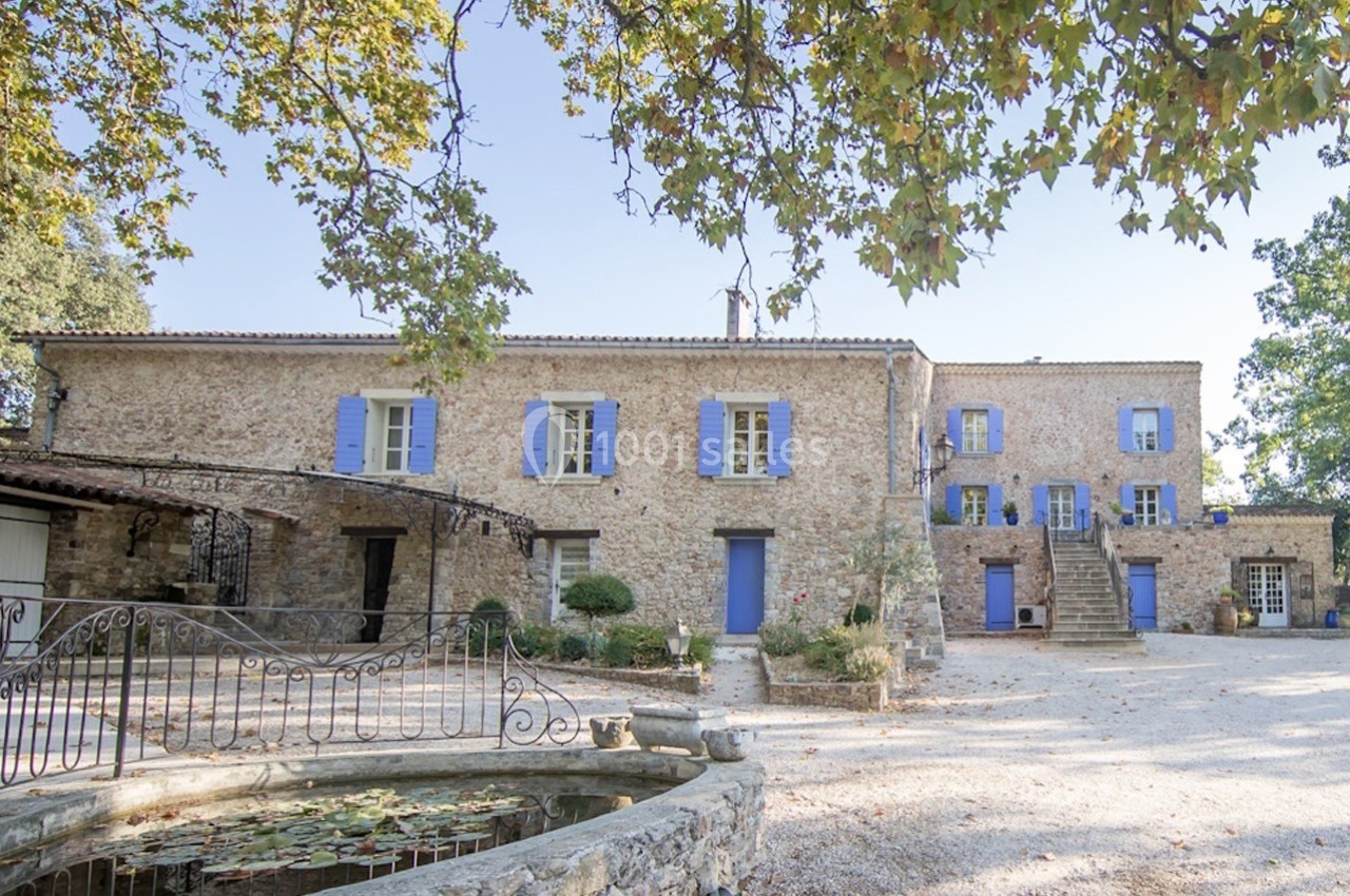 Façade d'une maison en pierre avec volets bleus, entourée d'arbres, avec une fontaine au premier plan.