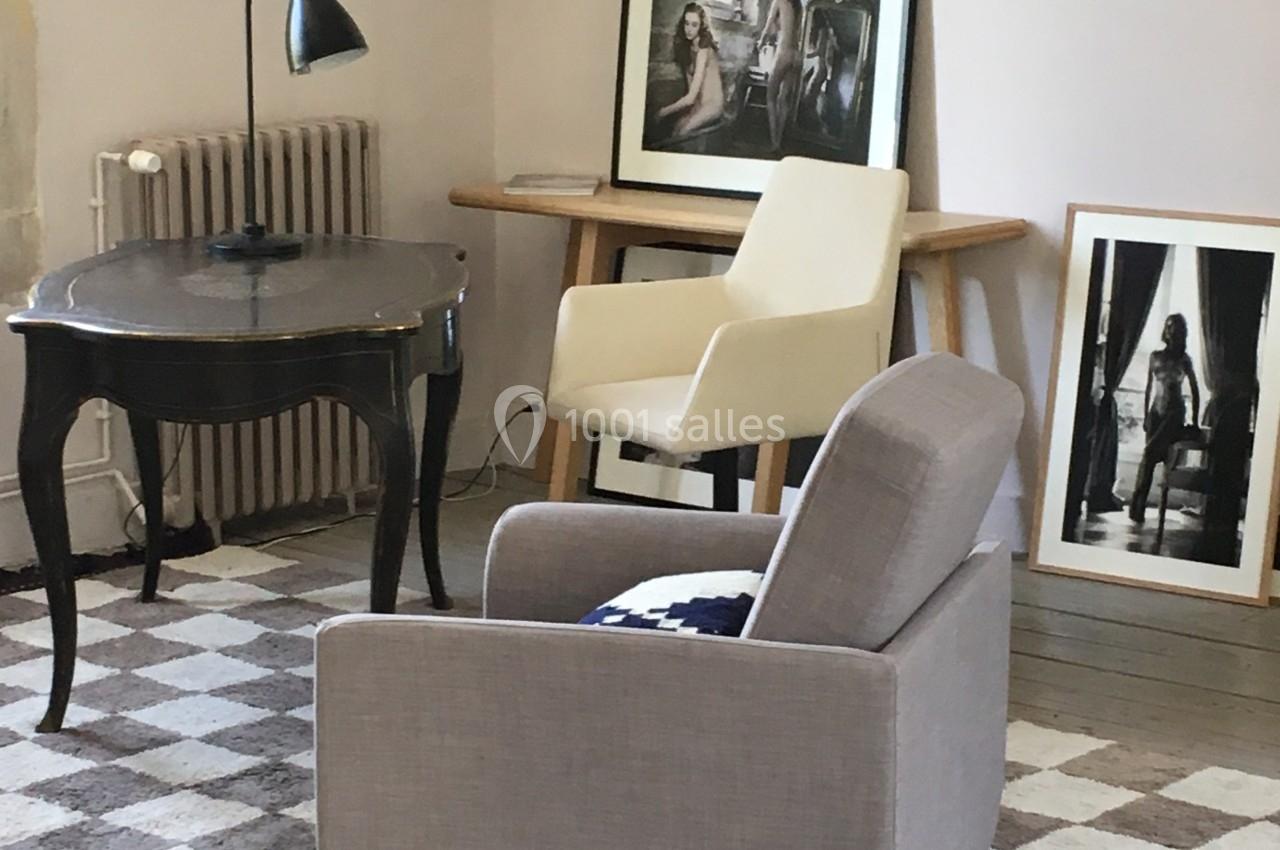Salon avec fauteuils, bureau noir, lampe de table et cadres posés sur un meuble en bois, tapis à motifs au sol.