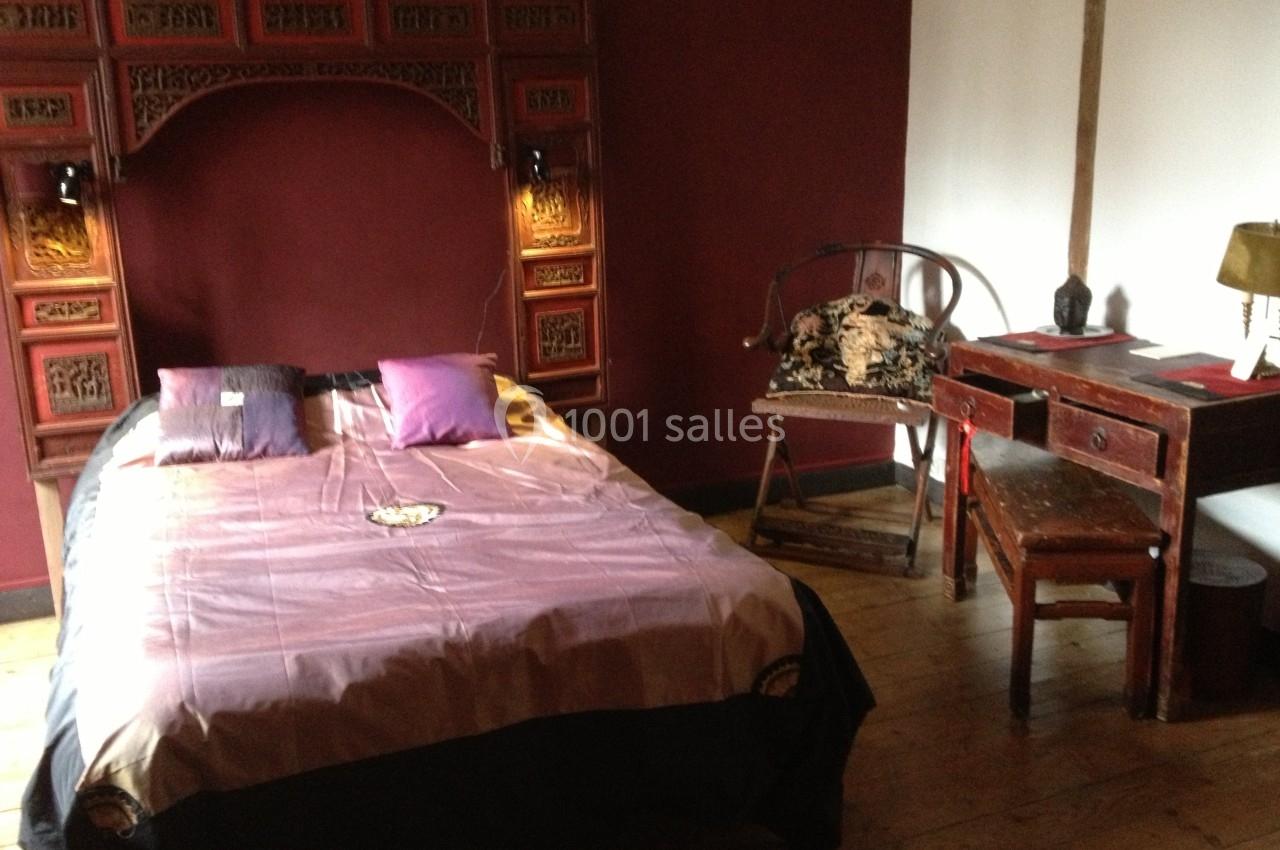 Chambre avec lit double, tête de lit sculptée en bois, bureau ancien, chaise et décoration dans des tons rouges et bois.