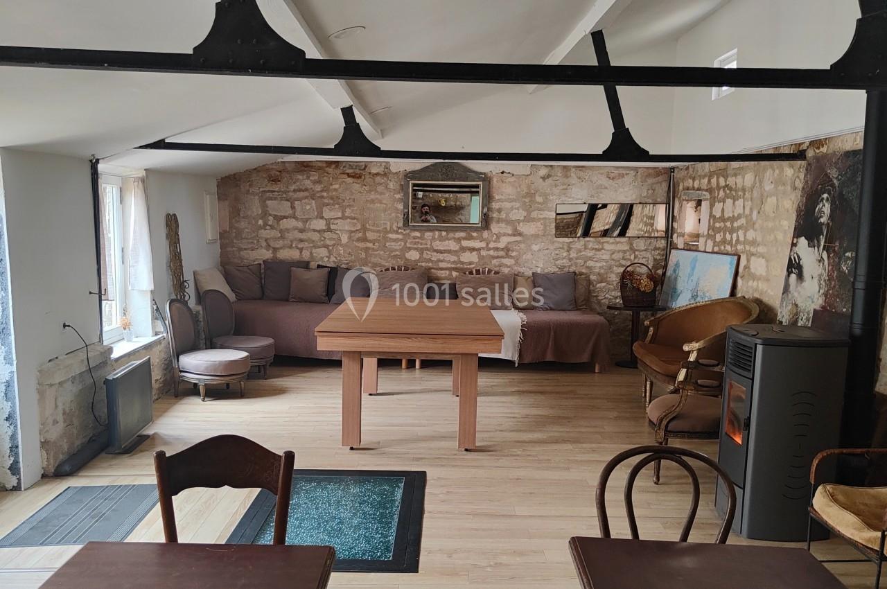 Salle lumineuse avec murs en pierre, tables en bois, poêle à bois et canapé au fond sous des poutres apparentes.