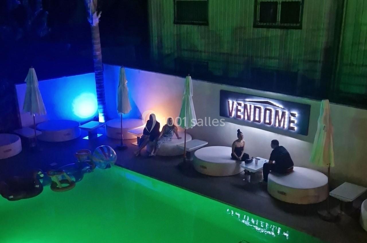 Personnes assises près d'une piscine éclairée, entourées de parasols fermés, avec une enseigne lumineuse ’Vendôme’.
