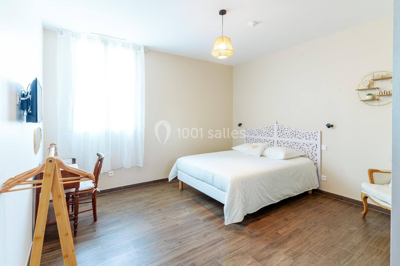 Chambre lumineuse avec un lit double, parquet au sol, mobilier en bois et une fenêtre avec rideaux blancs.