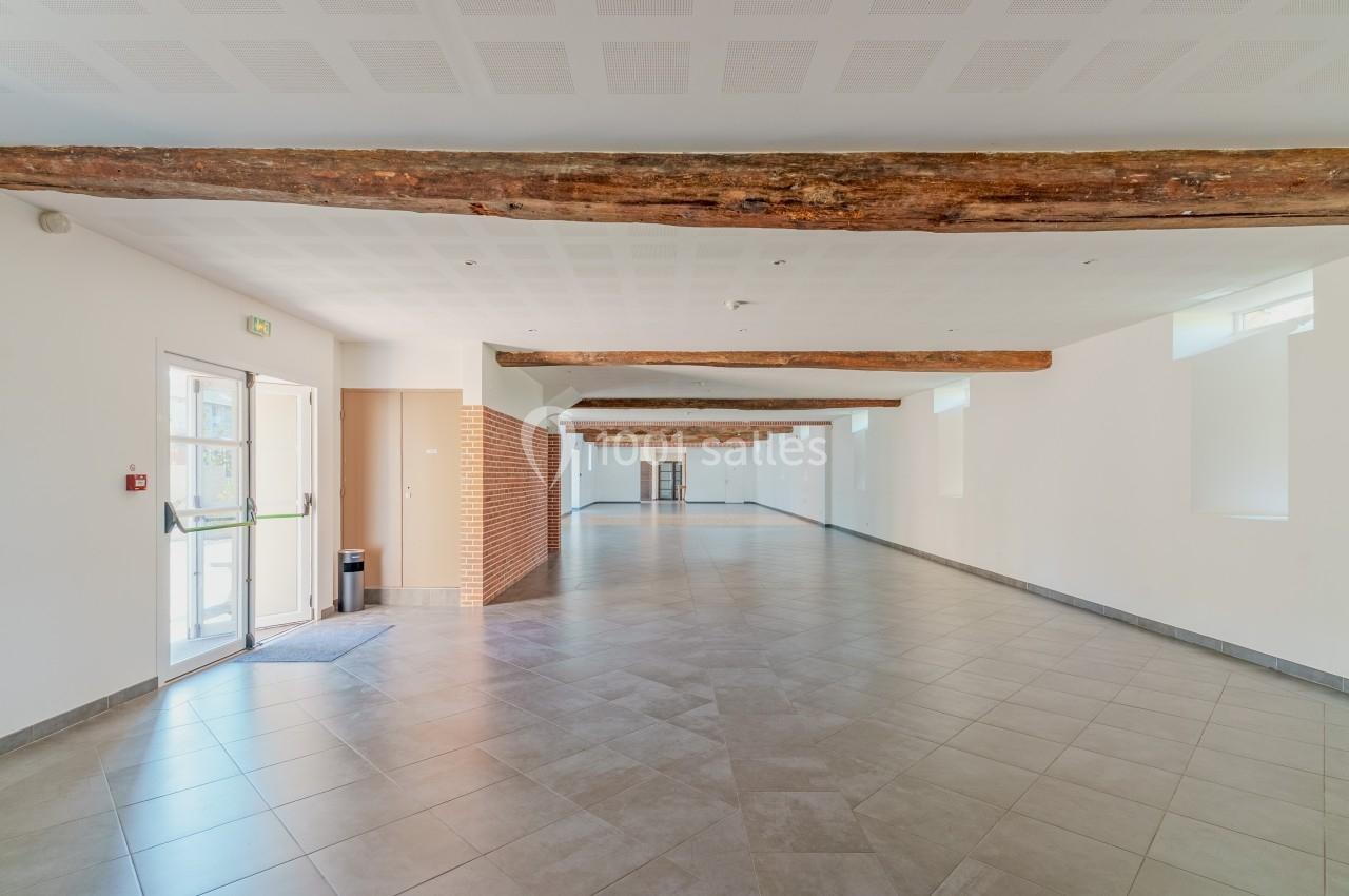 Salle vide avec carrelage au sol, poutres en bois au plafond, murs blancs et lumière naturelle entrant par des fenêtres.