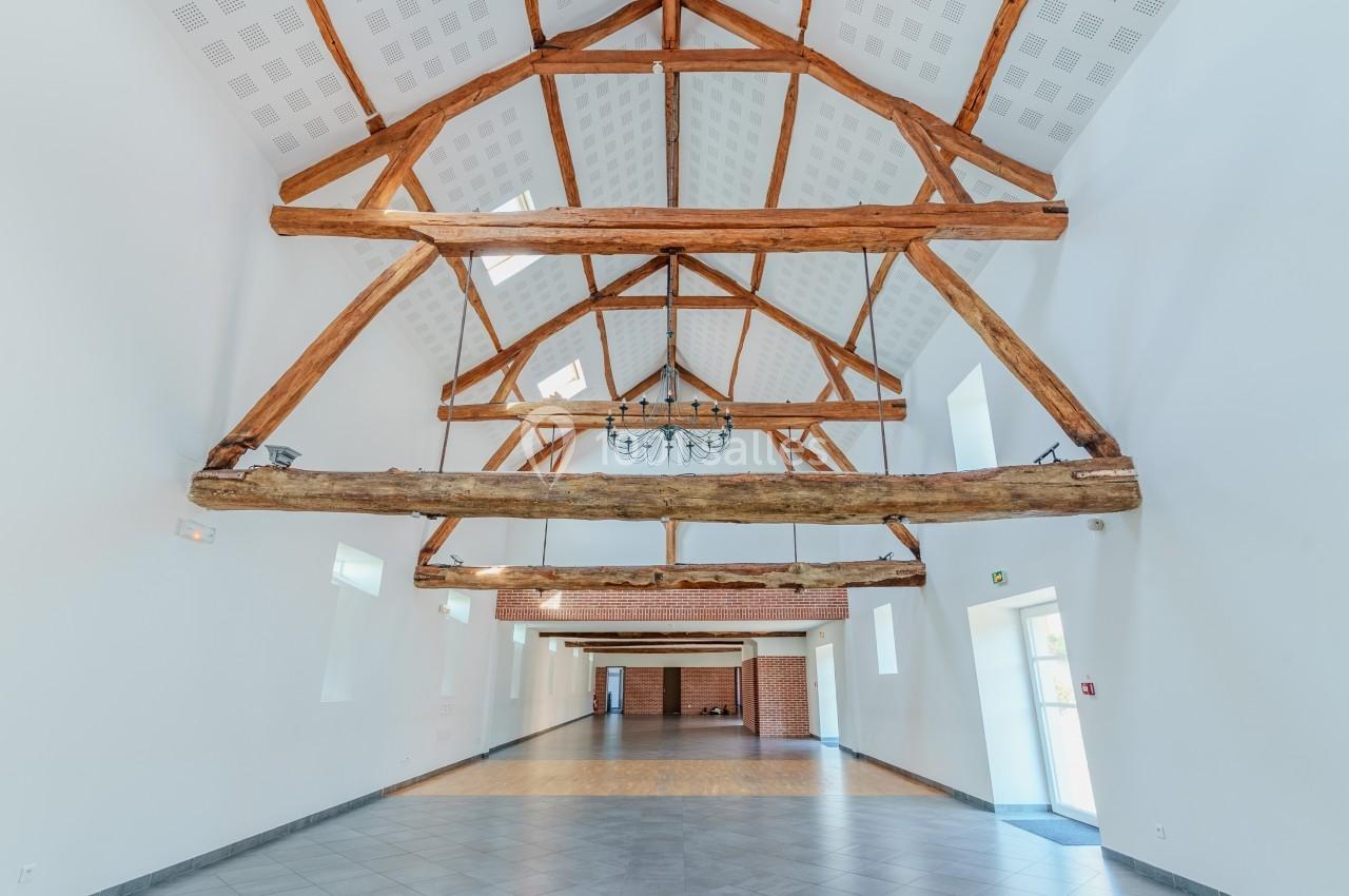 Charpente en bois apparente dans une grande salle lumineuse avec murs blancs et sol en carrelage gris.