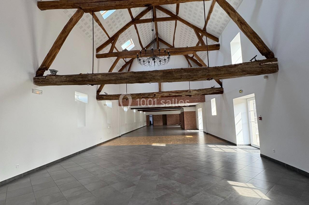 Grande salle lumineuse avec poutres apparentes en bois, sol en carrelage gris et murs blancs.