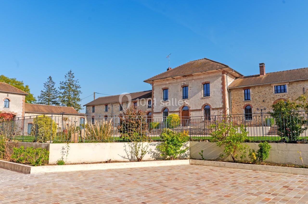 Bâtiment en pierre avec toit en tuiles, entouré d'un jardin et d'une terrasse pavée sous un ciel bleu.