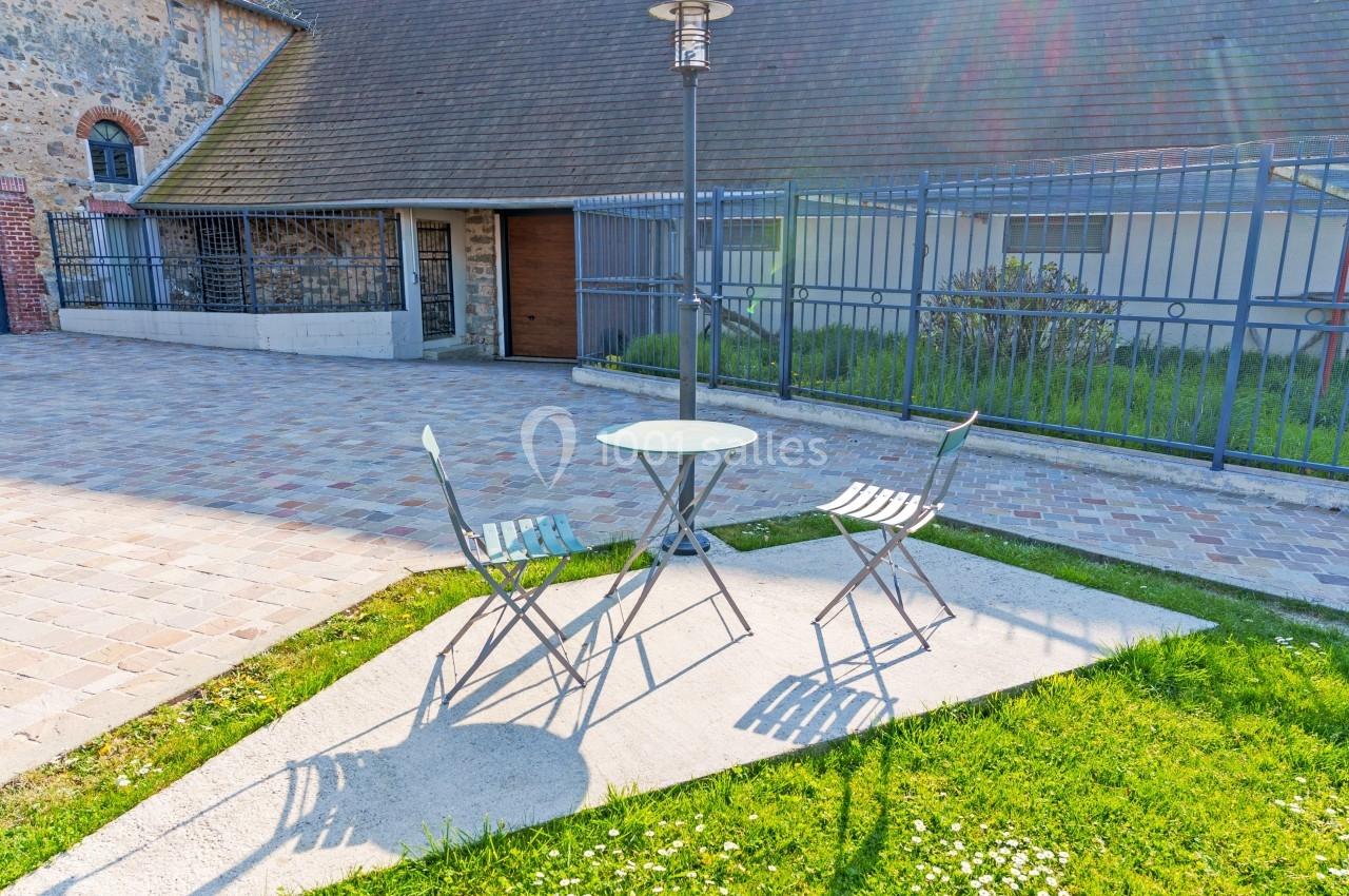 Deux chaises et une table ronde en métal sur une petite terrasse ensoleillée, près d'une maison en pierre.