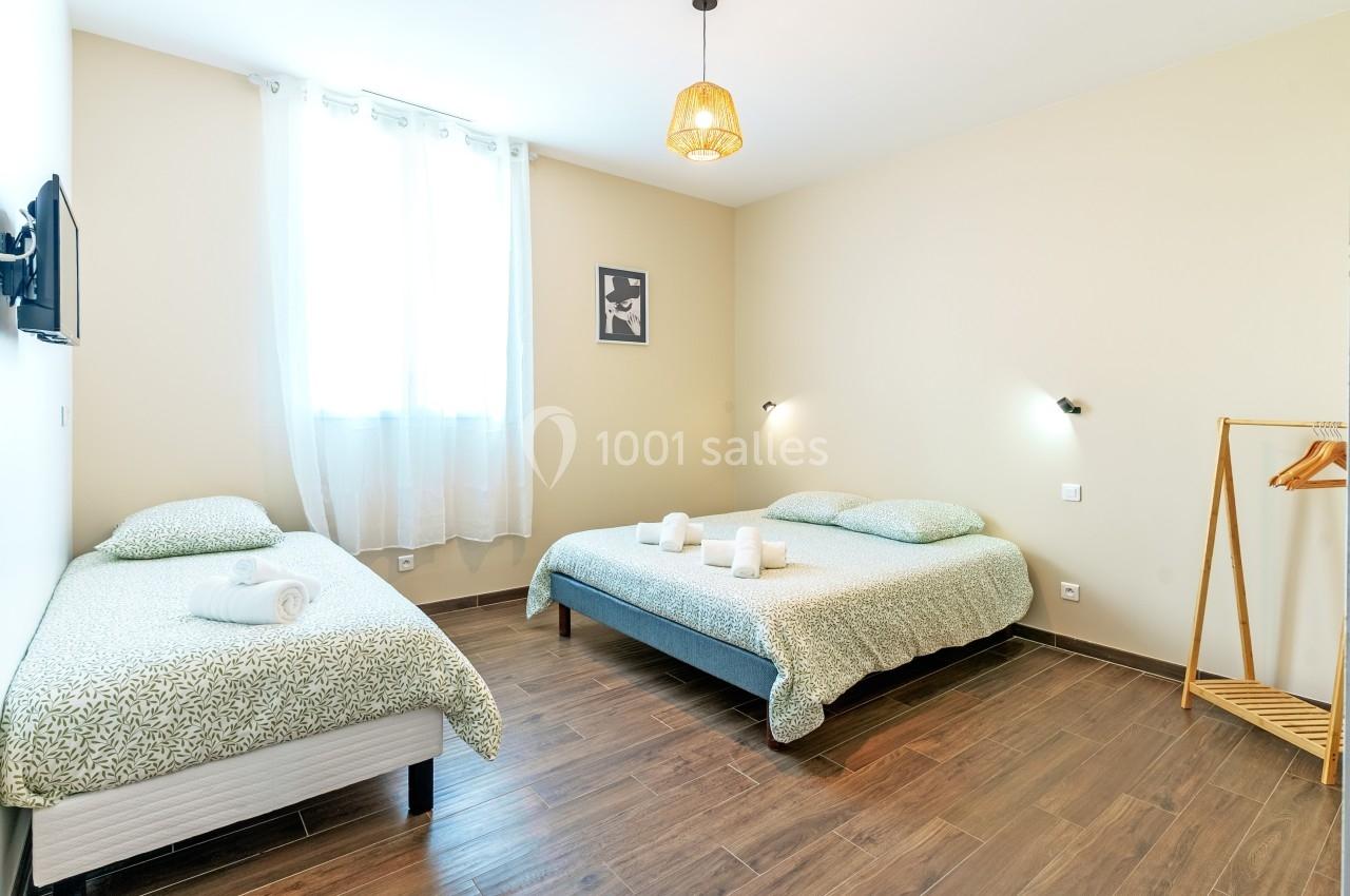 Chambre lumineuse avec un lit double, un lit simple, sol en bois, portant à vêtements et fenêtre avec rideaux blancs.