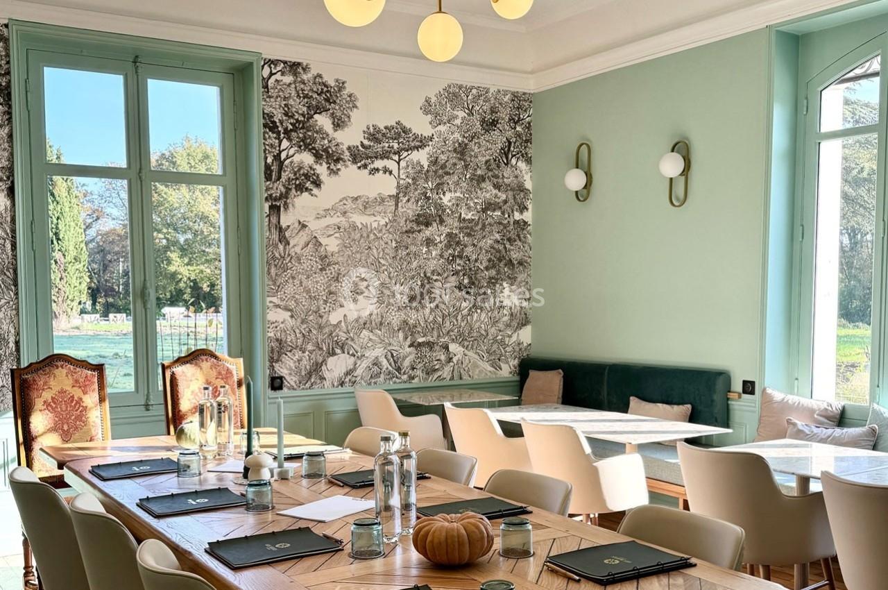 Salle lumineuse avec une grande table en bois, chaises modernes, fresque murale et vue sur un jardin à travers les fenêtres.