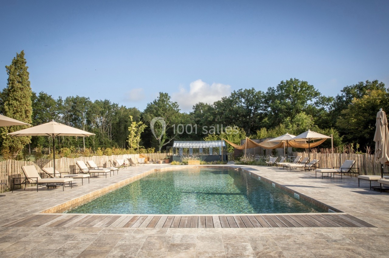 Piscine extérieure entourée de chaises longues, parasols et végétation, sous un ciel dégagé.