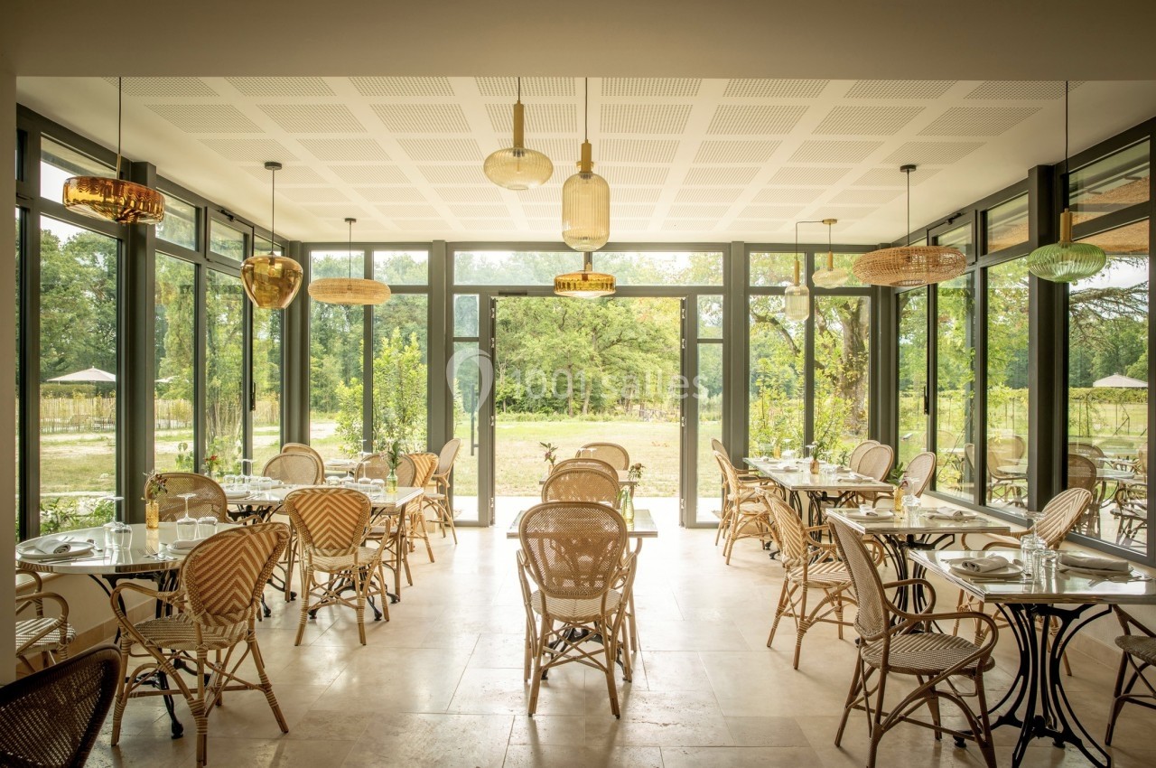 Salle lumineuse avec des tables et chaises en rotin, grandes baies vitrées donnant sur un jardin verdoyant.