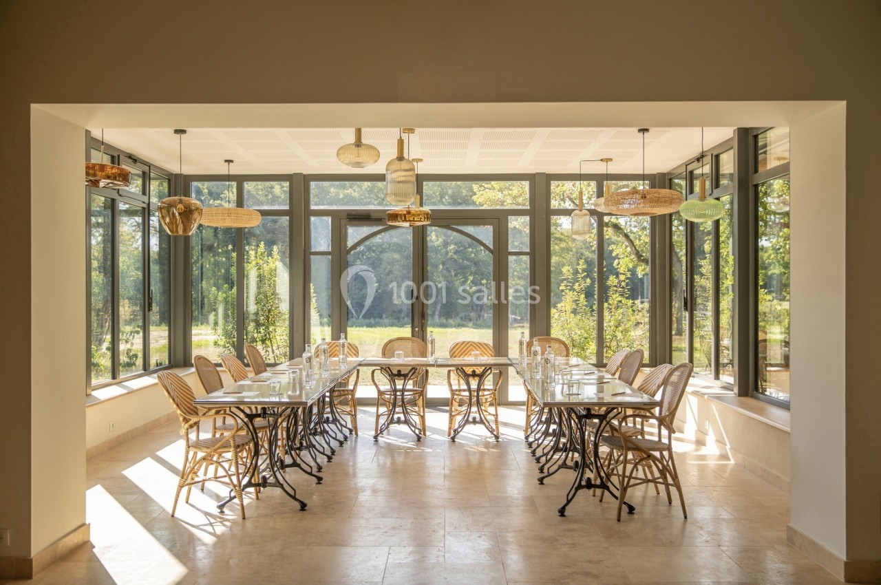 Salle lumineuse avec de grandes baies vitrées, table longue entourée de chaises en rotin et vue sur un jardin verdoyant.