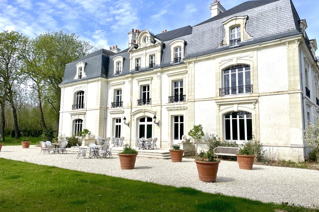 Façade d'un grand bâtiment blanc de style classique avec terrasse aménagée et pots de fleurs, entouré de verdure.