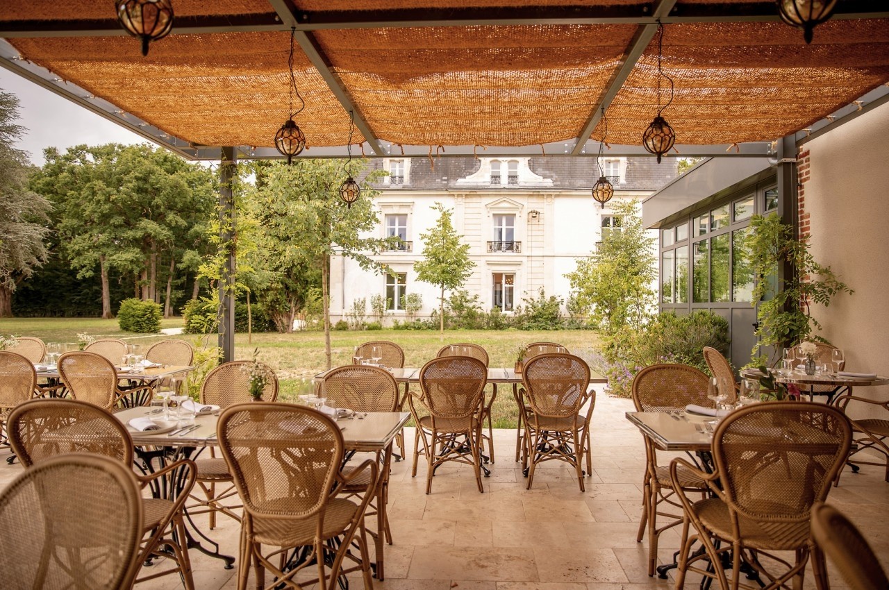 Terrasse d'un restaurant avec des tables et chaises en rotin, vue sur un jardin et une bâtisse élégante en arrière-plan.