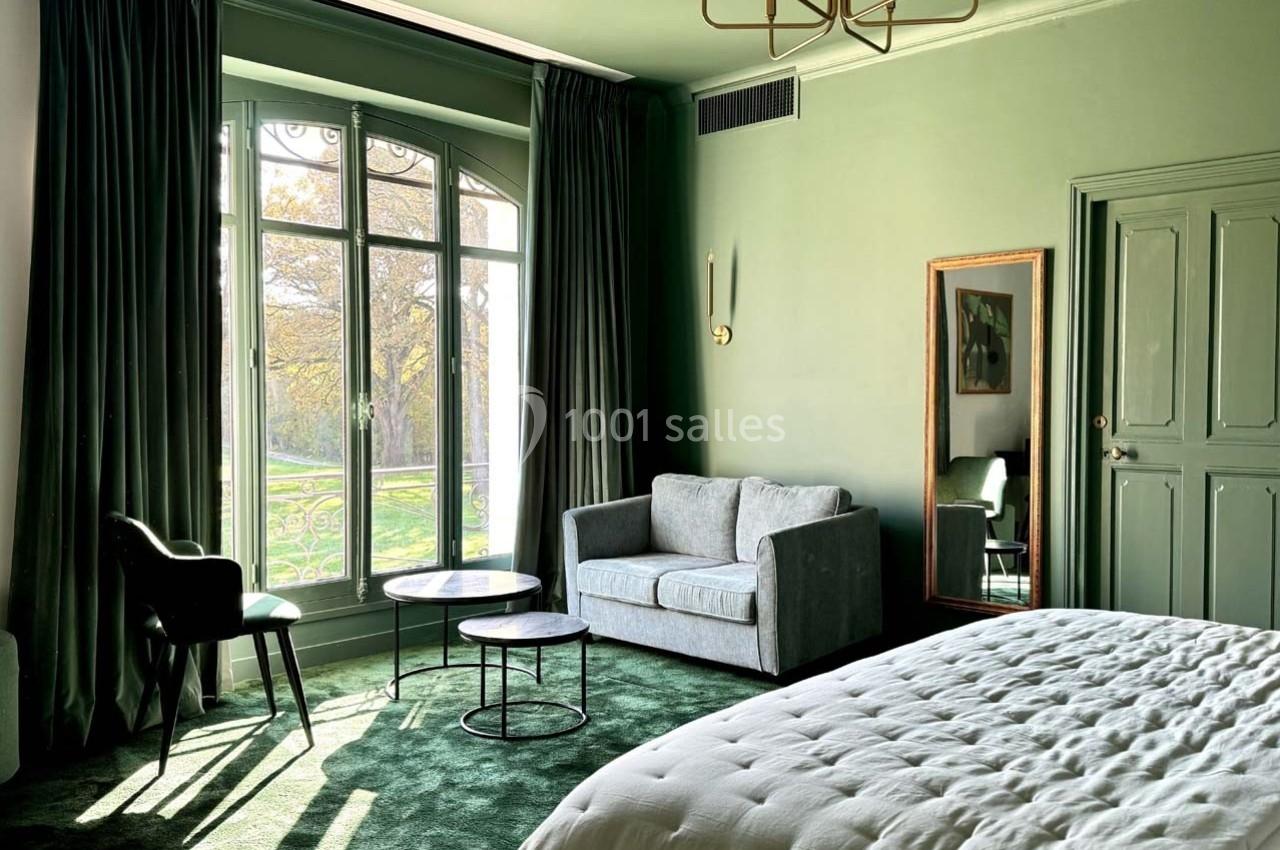 Chambre lumineuse avec murs verts, lit, canapé gris, tables basses, chaise noire et grande fenêtre donnant sur un jardin.