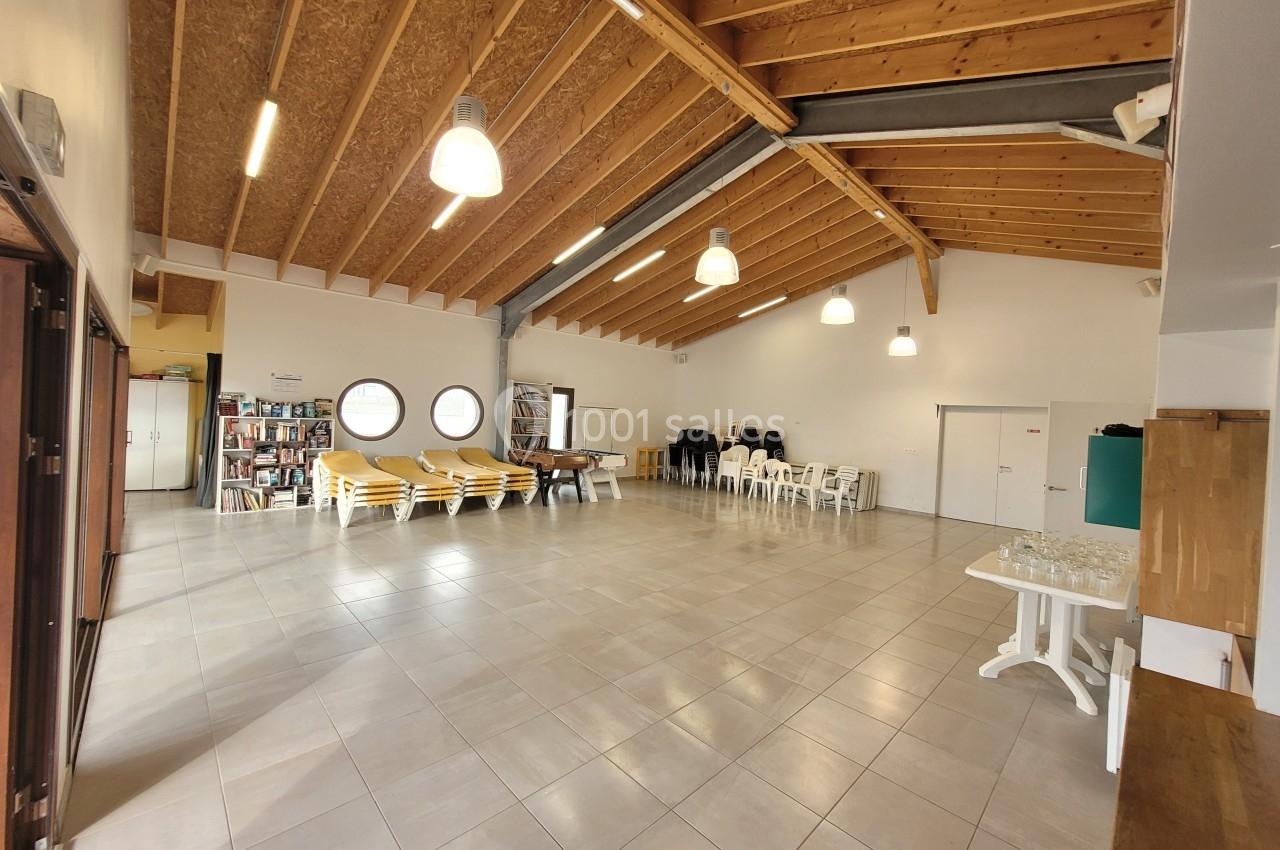 Salle spacieuse avec plafond en bois, tables, chaises empilées, étagères de livres et grandes fenêtres rondes.