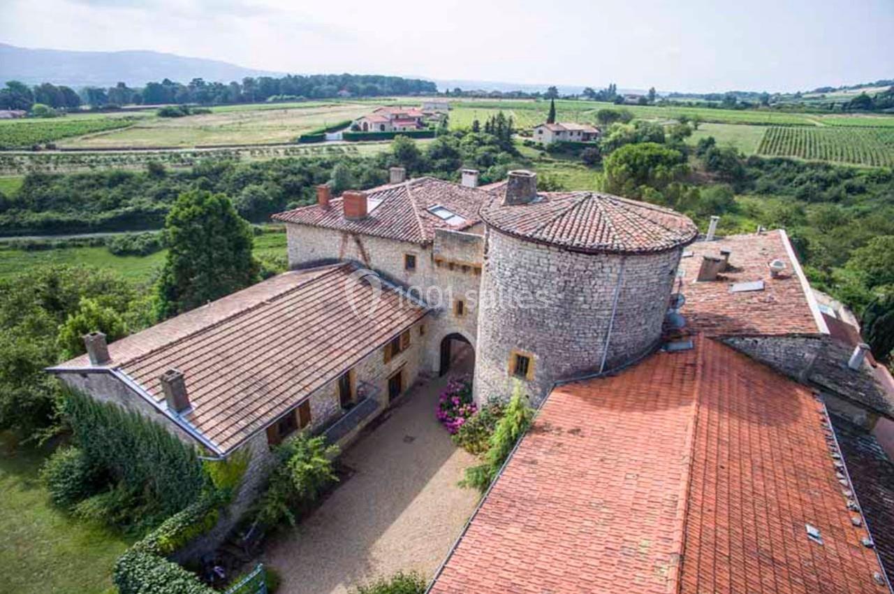 Vue aérienne d'un bâtiment en pierre avec des toits en tuiles rouges, entouré de champs et de verdure.