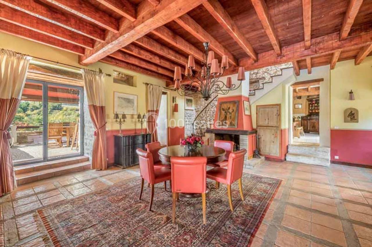Salle à manger rustique avec poutres apparentes, table ronde entourée de chaises rouges et vue sur une terrasse extérieure.