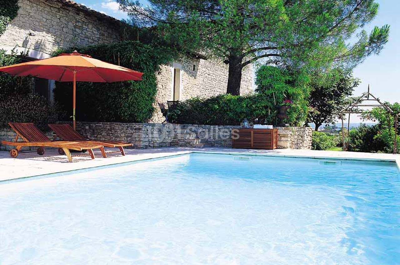 Piscine extérieure entourée de murs en pierre, transats en bois, parasol orange et végétation méditerranéenne.