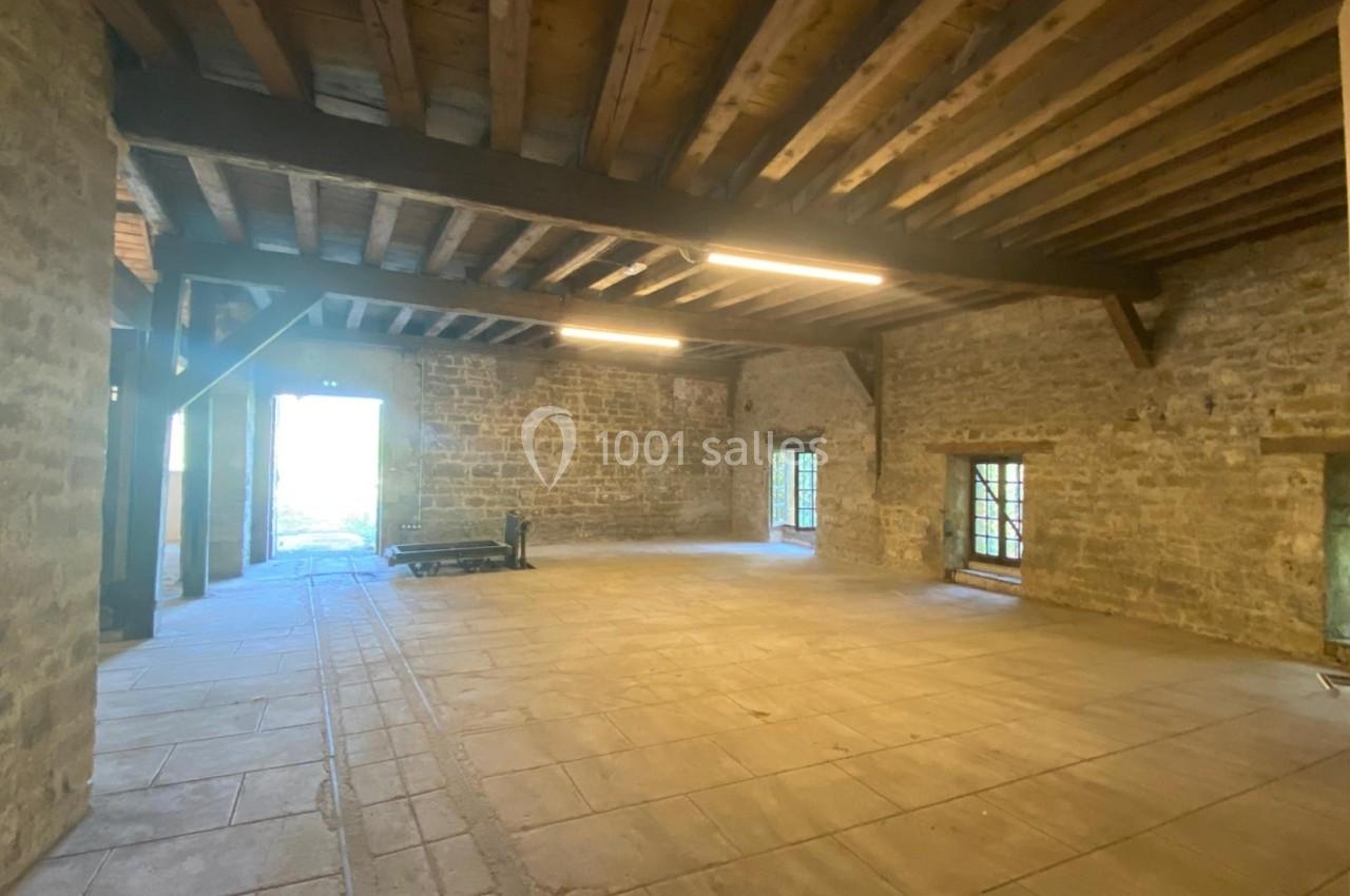 Intérieur d'une grande pièce en pierre avec plafond en poutres apparentes, sol en dalles et lumière naturelle entrant par…
