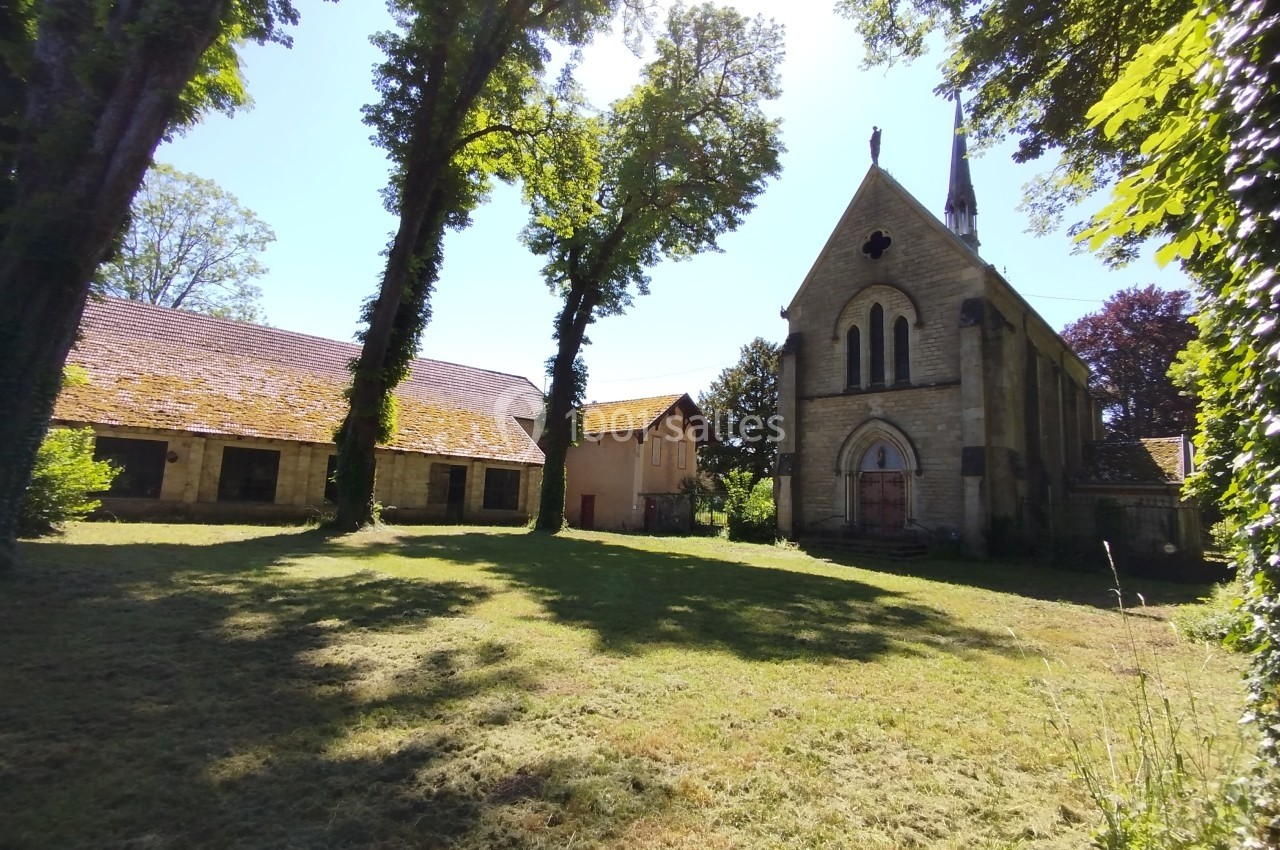 Vue d'une petite église en pierre entourée d'arbres, avec des bâtiments annexes et une pelouse ensoleillée.
