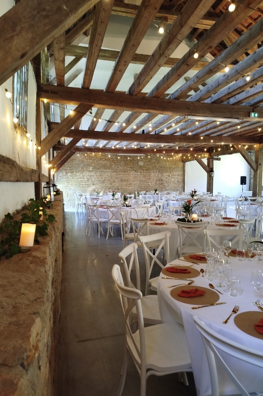 Salle de réception décorée avec des tables rondes dressées, des chaises blanches et des guirlandes lumineuses suspendues.