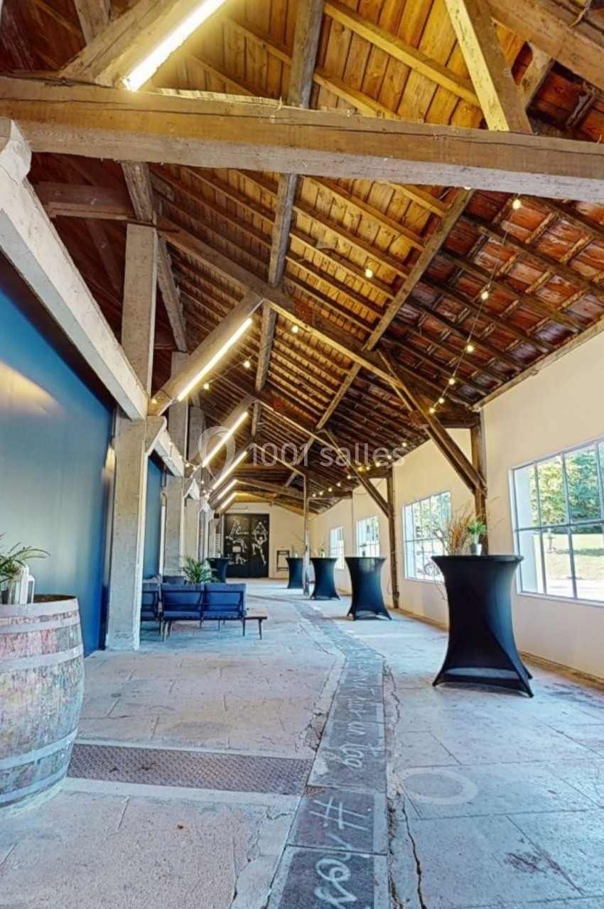 Intérieur d'une salle avec charpente en bois, tables hautes, tonneau décoratif et grandes fenêtres laissant entrer la…