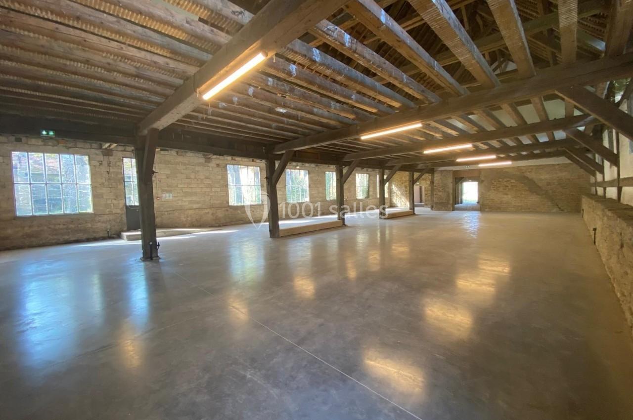 Intérieur d'un grand espace industriel rénové avec poutres en bois apparentes, murs en pierre et sol en béton poli.