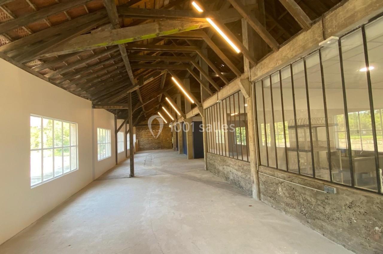 Intérieur d'un bâtiment rénové avec charpente en bois apparente, grandes fenêtres et murs en pierre et verre.
