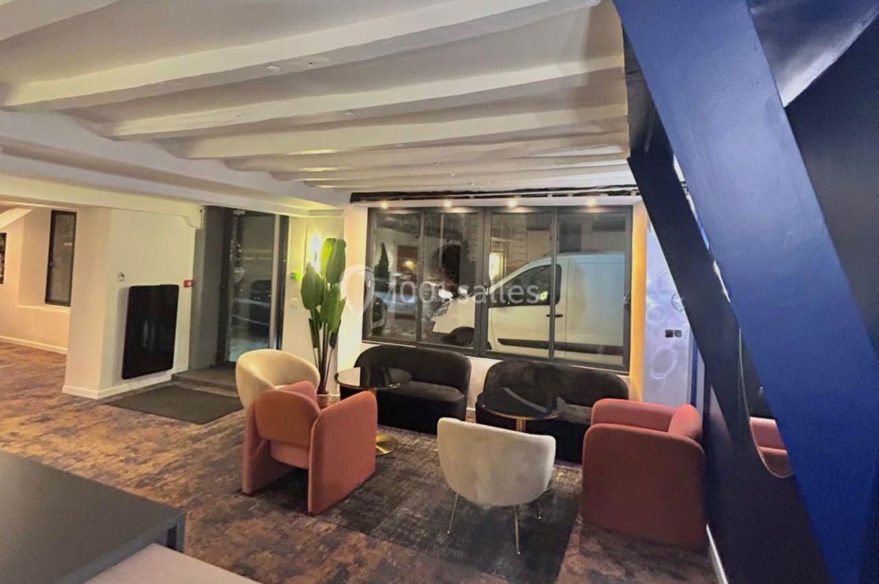 Espace intérieur lumineux avec fauteuils colorés, grande fenêtre donnant sur la rue et poutres apparentes au plafond.