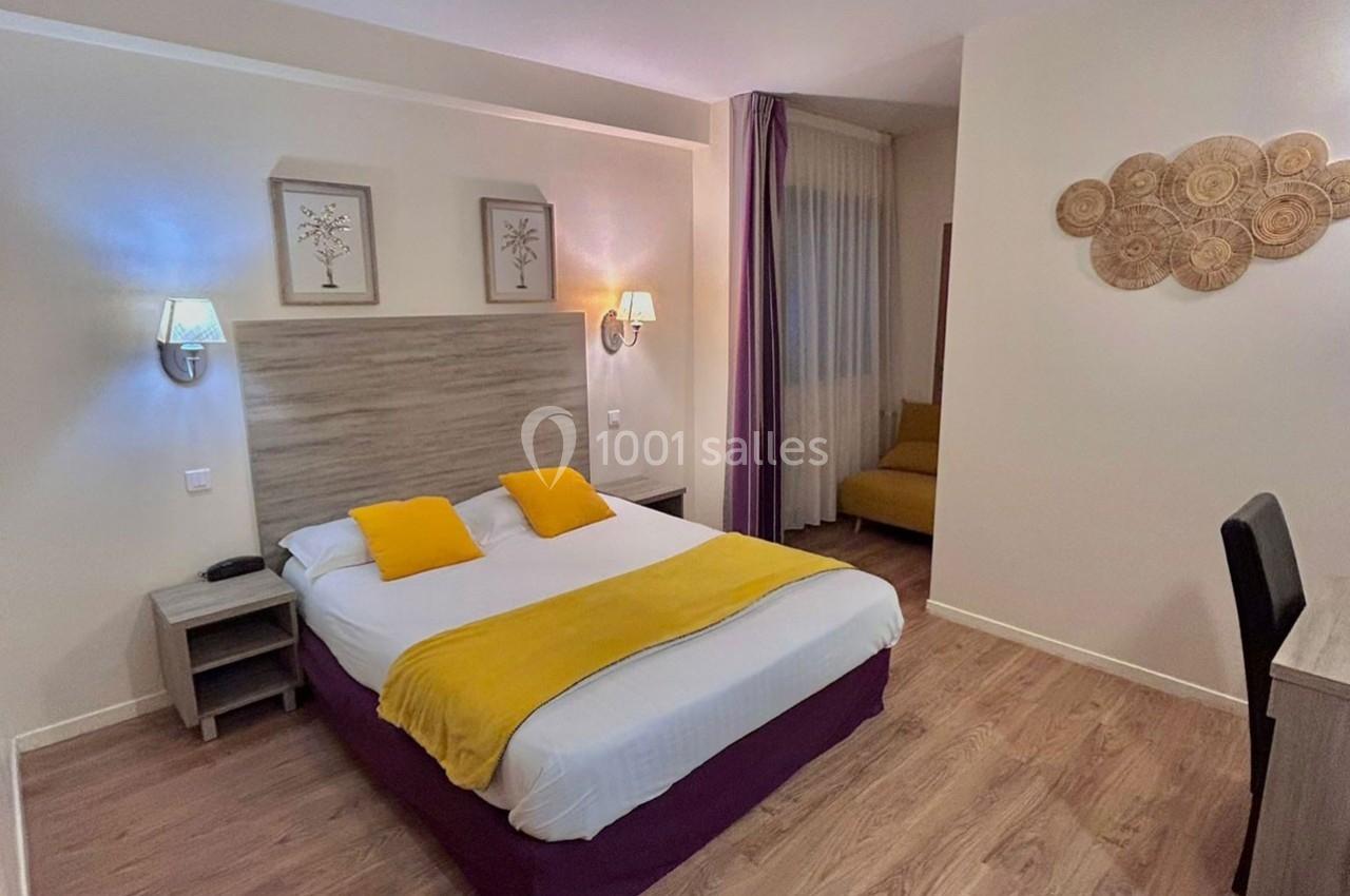 Chambre lumineuse avec un lit double, coussins jaunes, décoration murale et coin salon visible en arrière-plan.