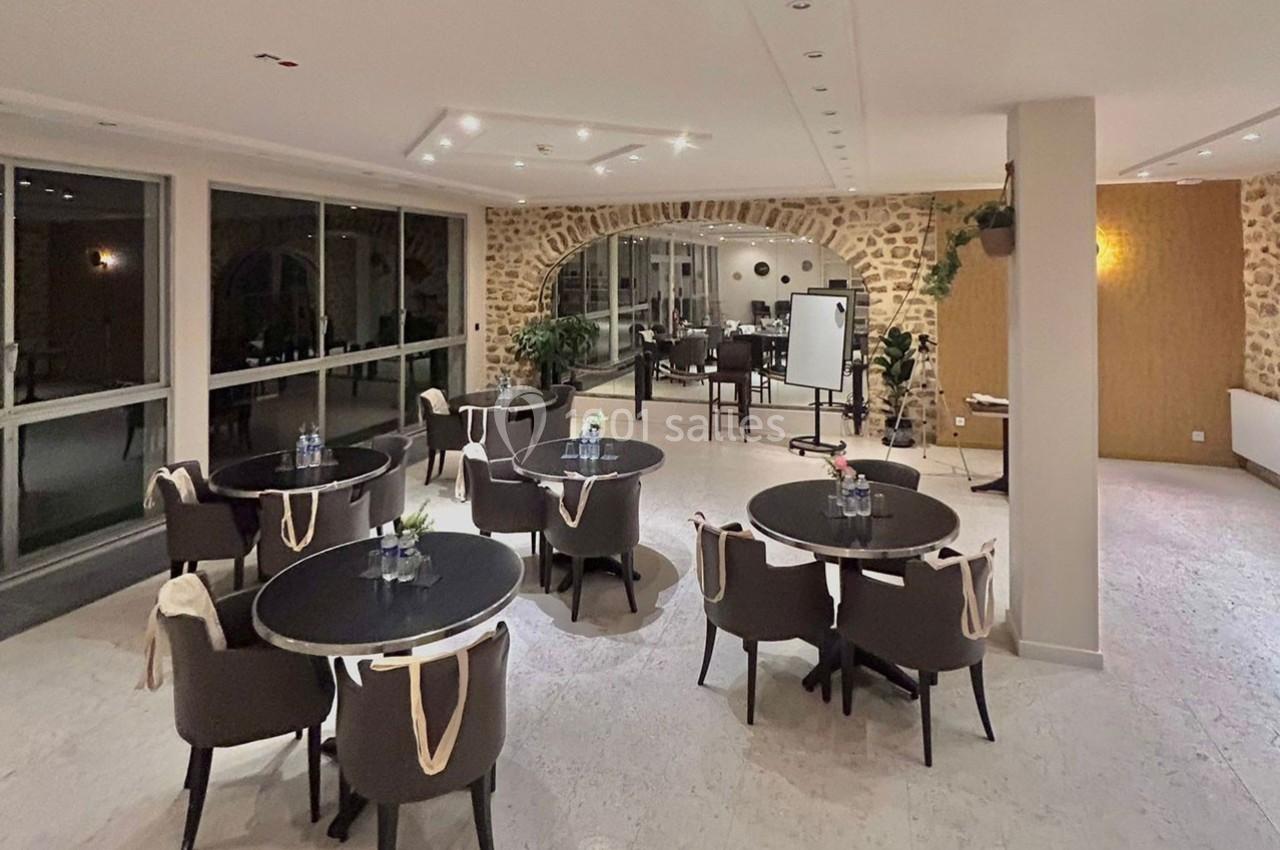 Salle lumineuse avec tables rondes, chaises rembourrées, murs en pierre et grandes baies vitrées donnant sur l'extérieur.