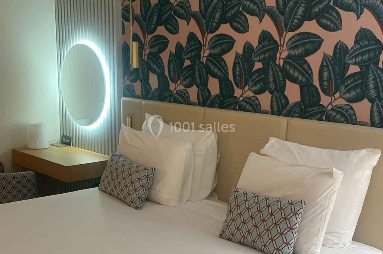 Lit double avec coussins décoratifs, table de chevet, miroir rond lumineux et mur tapissé de motifs végétaux.