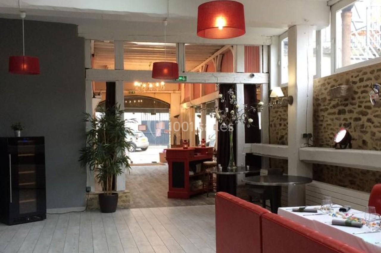 Salle lumineuse avec murs en pierre, mobilier moderne, lampes rouges suspendues et vue sur une entrée vitrée.