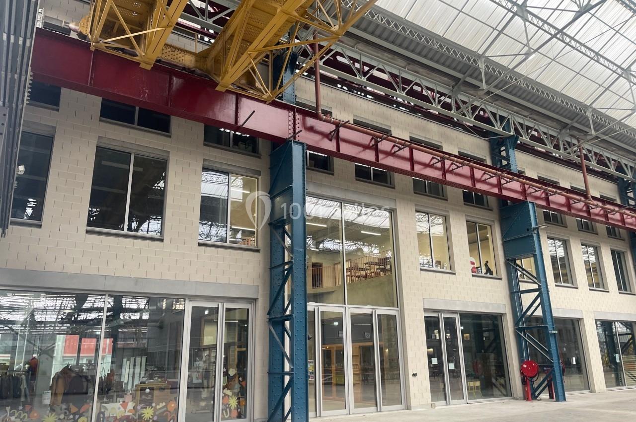 Structure industrielle rénovée avec de grandes baies vitrées, poutres métalliques colorées et plafond en verrière.