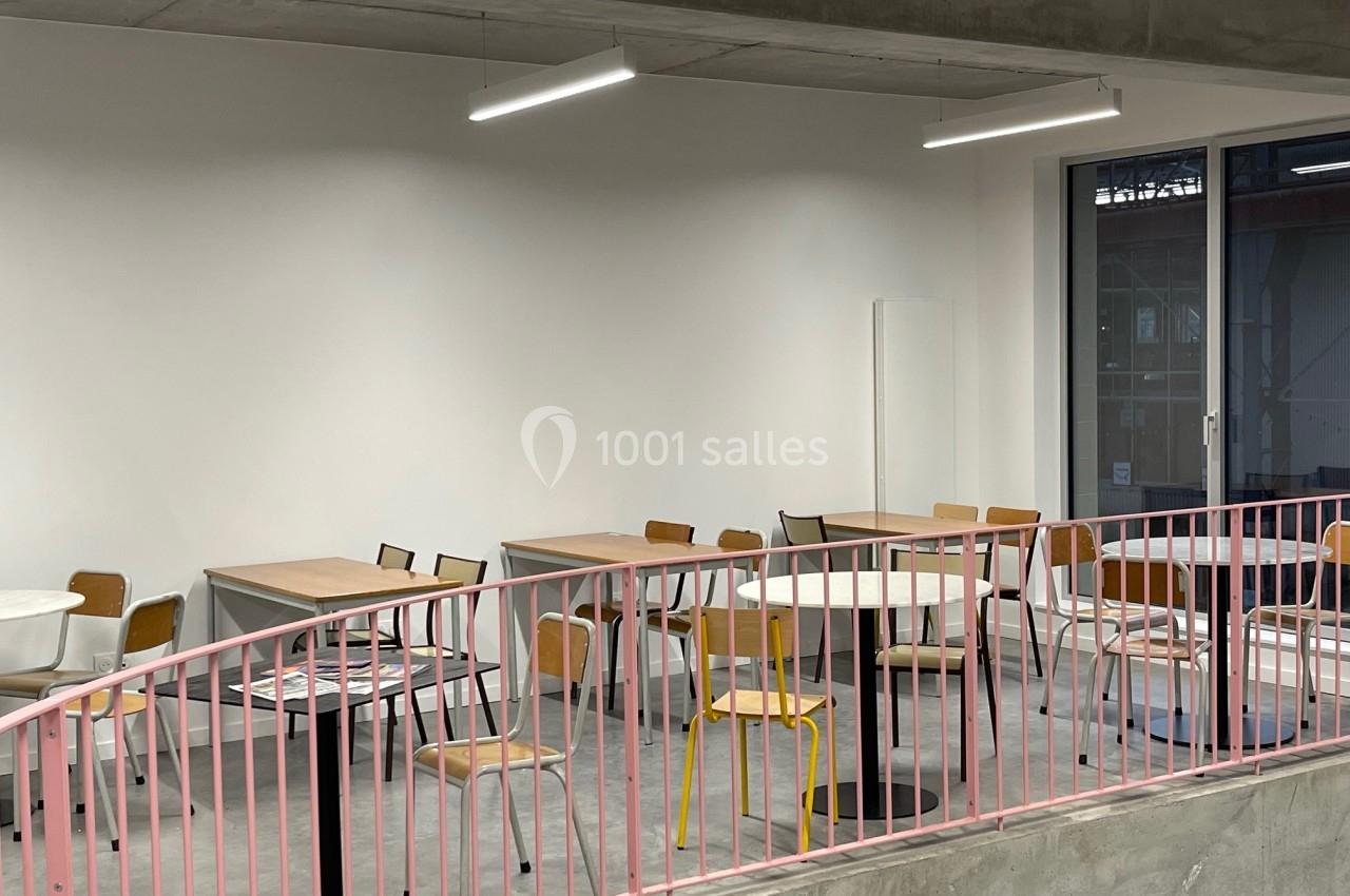 Salle lumineuse avec des tables et des chaises disposées de manière espacée, entourée d'une rambarde rose.