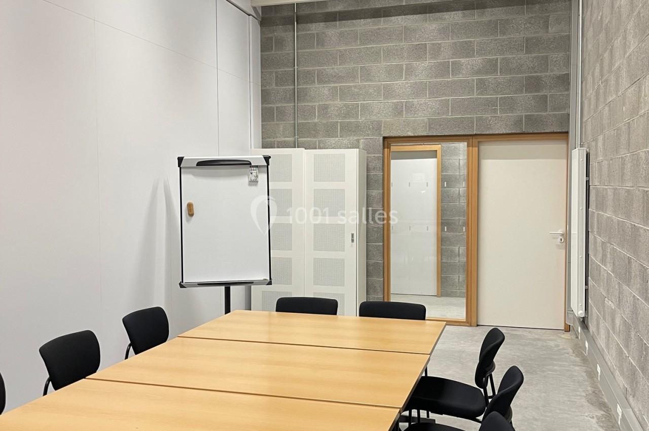 Salle de réunion avec table en bois, chaises noires, tableau blanc et murs en béton et panneaux blancs.