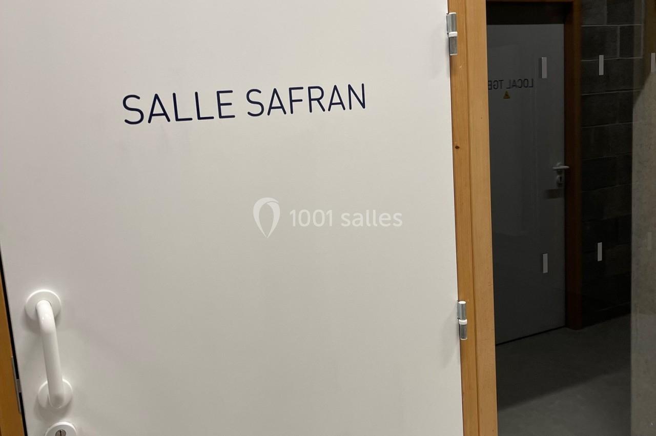 Porte blanche avec l'inscription ’Salle Safran’, encadrée de bois, donnant sur un espace intérieur en béton.