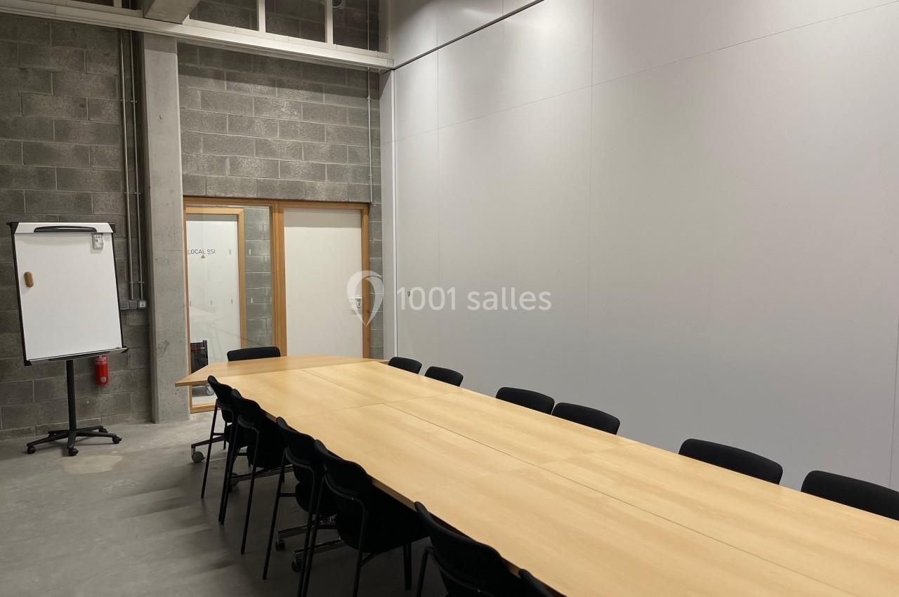 Salle de réunion avec une longue table en bois entourée de chaises noires, un tableau mobile et un mur en béton apparent.