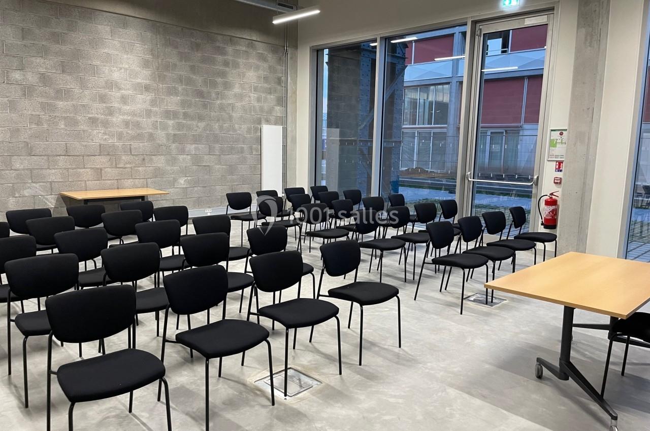 Salle de réunion avec des rangées de chaises noires vides, une table en bois et de grandes fenêtres donnant sur l'extérieur.